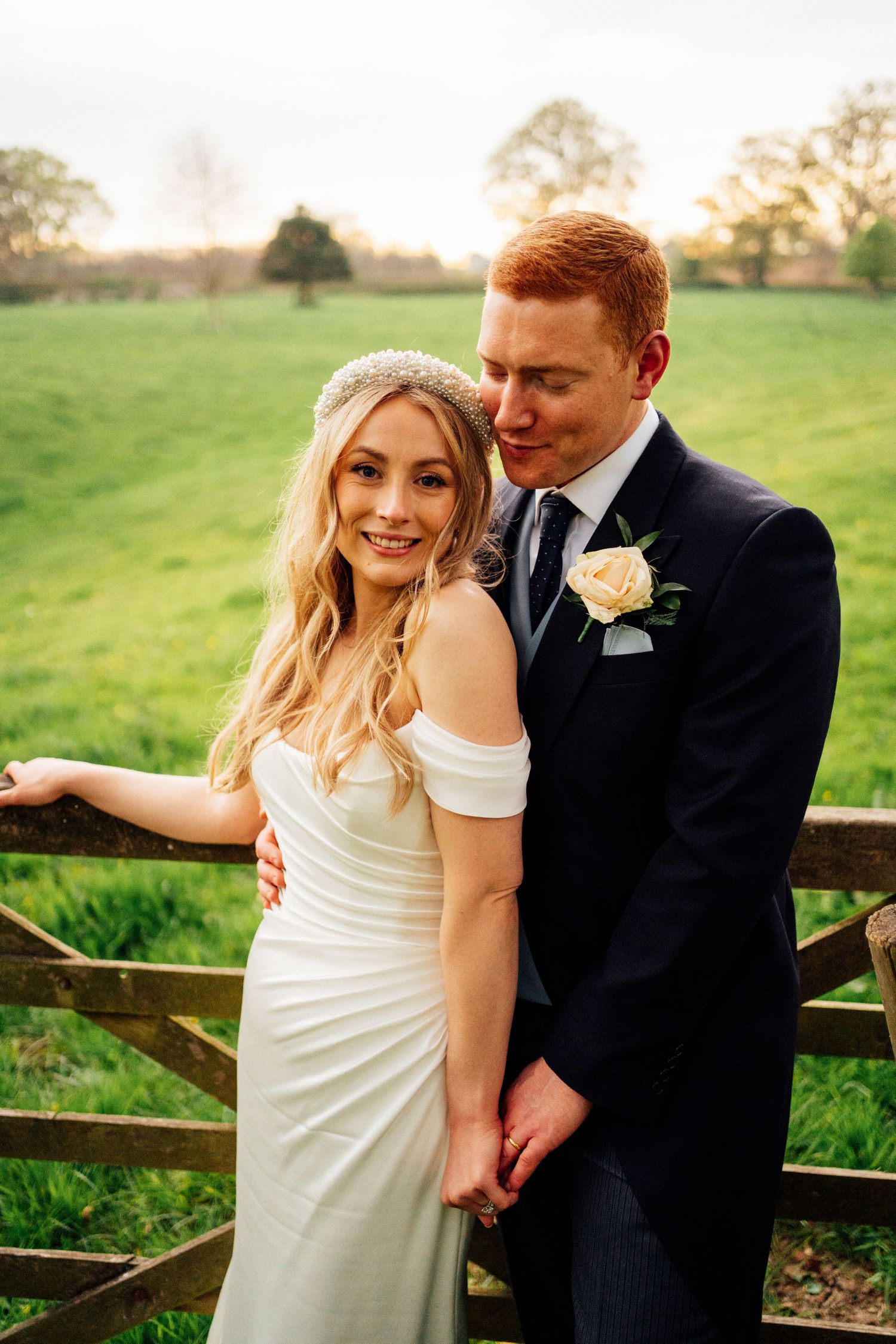 Romantic portrait of newlyweds posing by a wooden gate in a countryside setting.