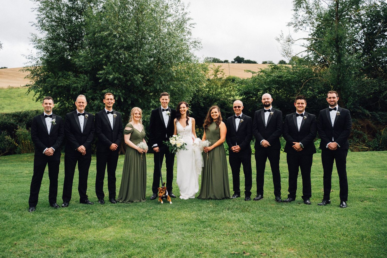 Wedding party in formal attire poses for group photos on a grassy lawn.