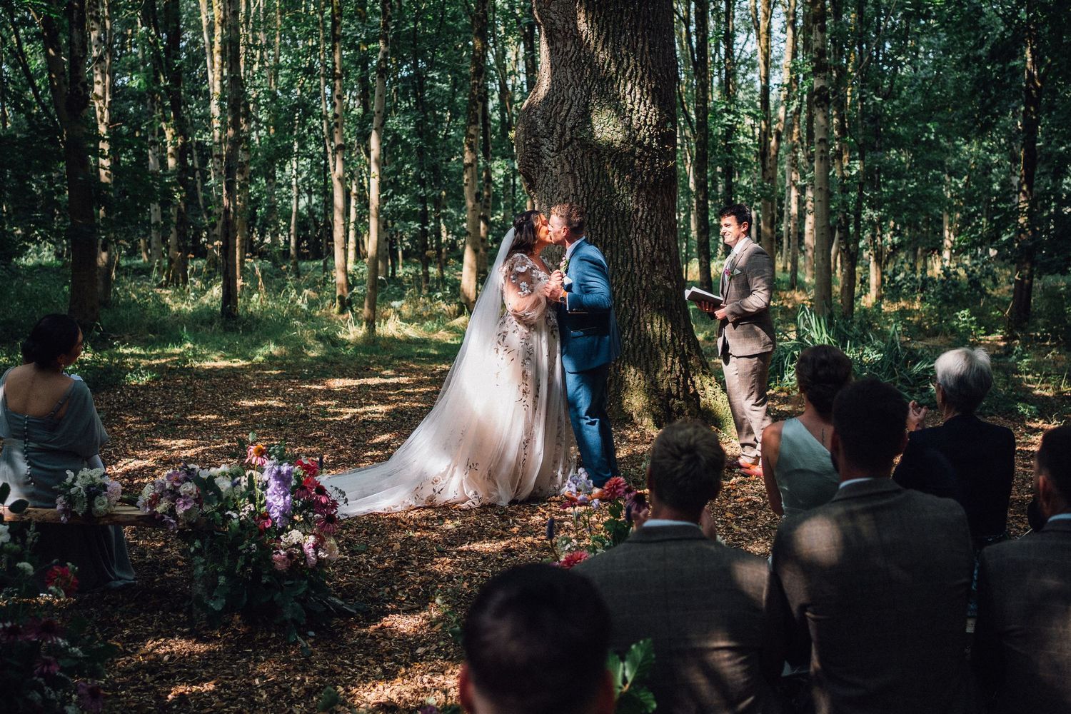 Guests observe a wedding ceremony in a peaceful woodland setting with dappled sunlight.