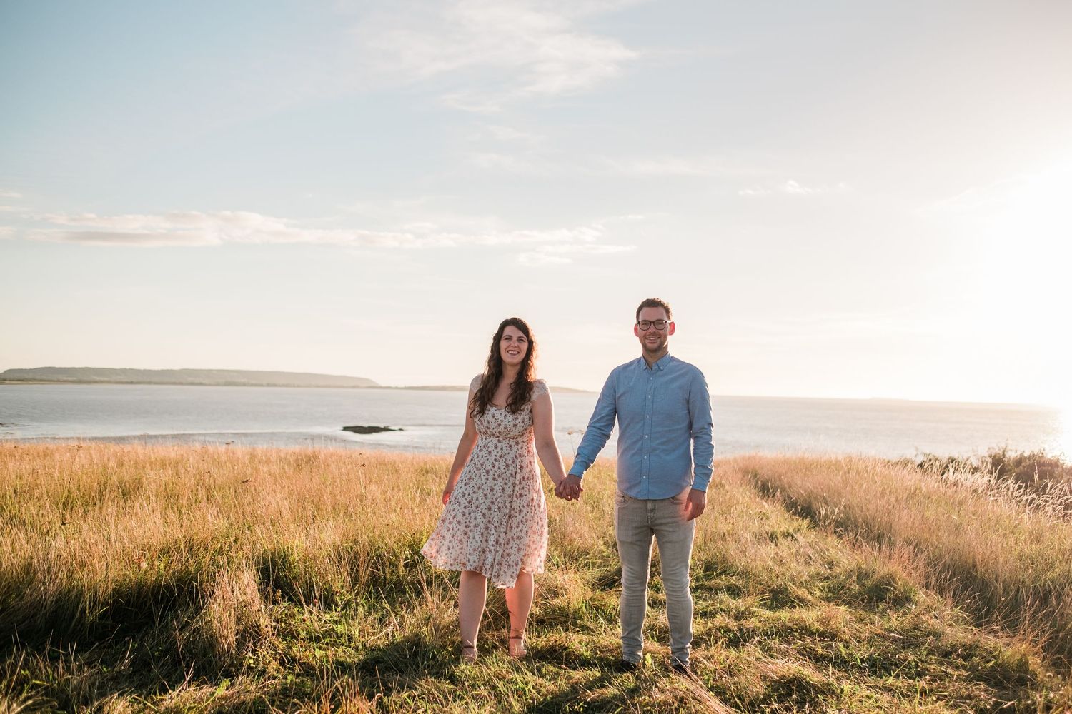 A couple walks hand in hand through a golden meadow at sunset near the ocean.