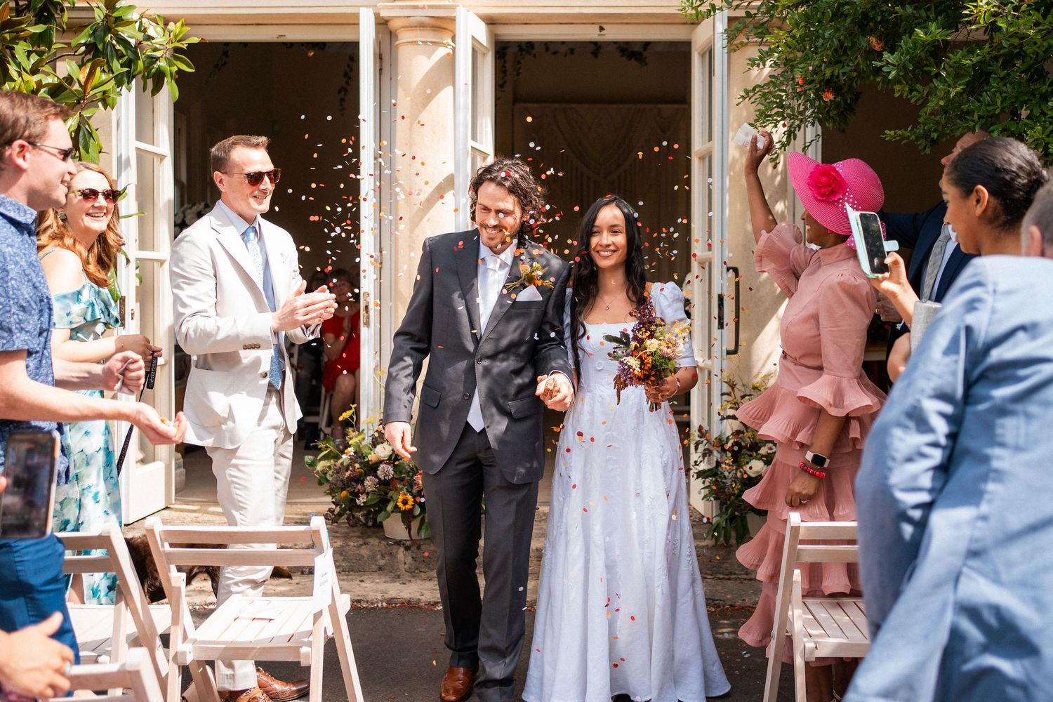 Bride and groom walk through confetti shower after outdoor wedding ceremony, surrounded by cheering guests.