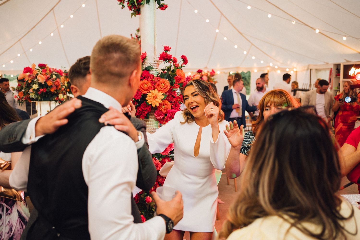 Wedding reception under a white tent with string lights and red floral decorations.