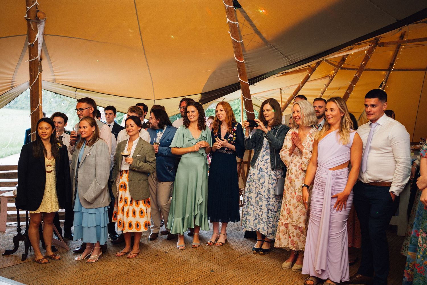 A group of guests in colorful summer dresses and formal attire gather under a tent at an outdoor celebration.