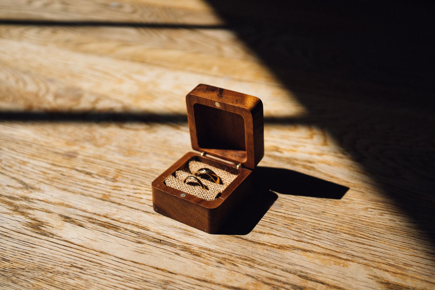 A small wooden ring box with wedding bands inside sits on a rustic wooden surface in dramatic lighting.