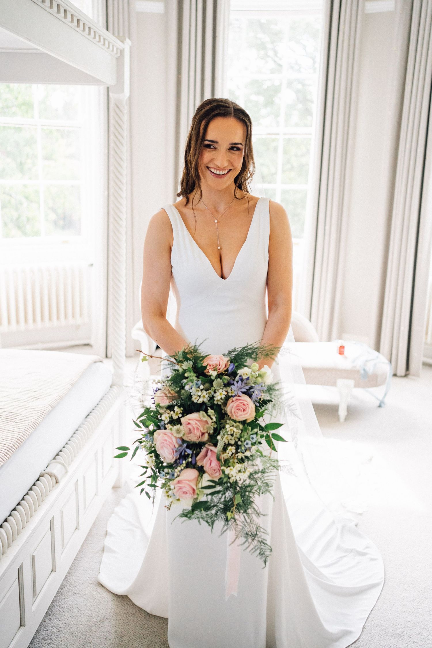 A bride in a classic white V-neck wedding gown holds a romantic bouquet of pink roses and greenery in a bright room at Eastington Park