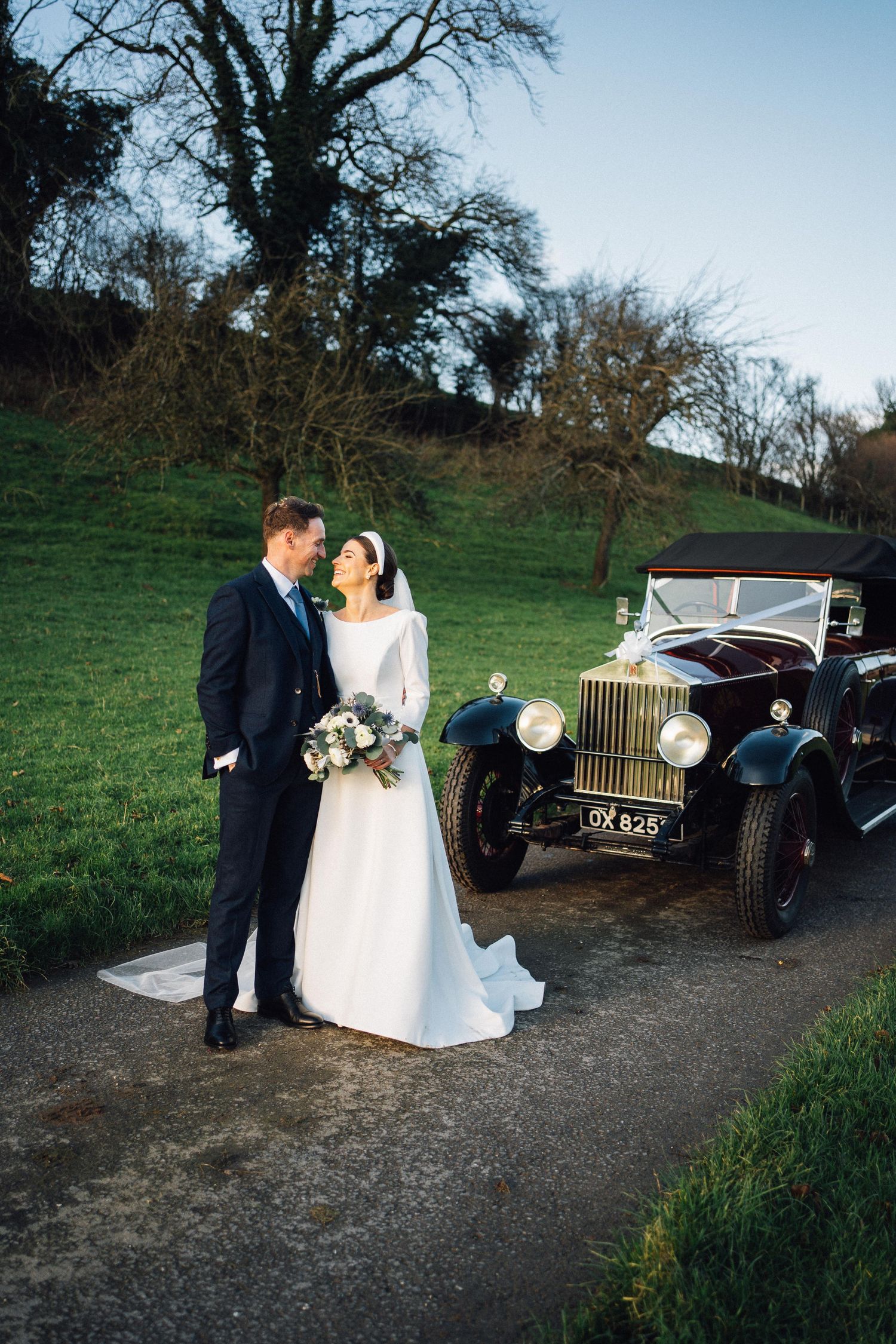 A romantic wedding couple poses with vintage cars in a rustic outdoor setting as the sun sets behind bare trees.