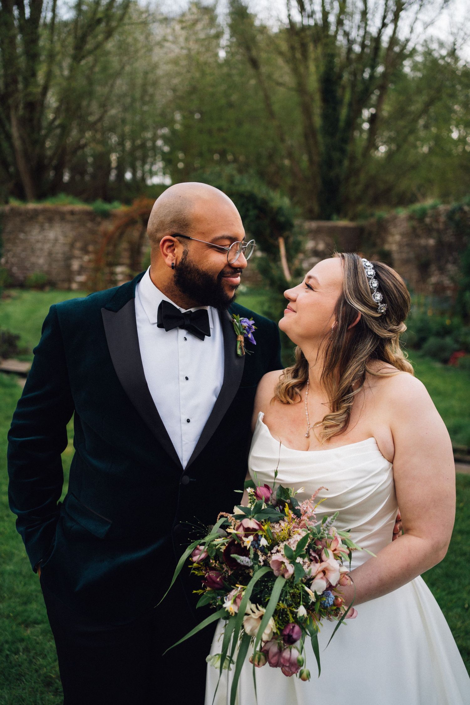 A happy couple shares romantic moments in a garden setting while wearing formal wedding attire and holding a floral bouquet.