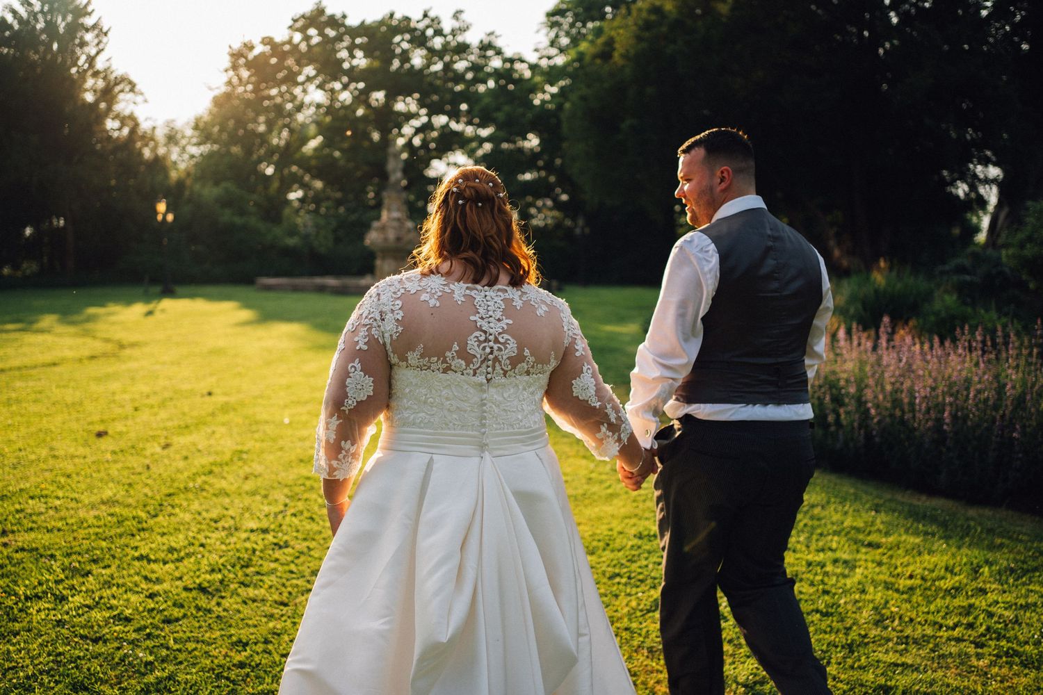 Newlyweds walk hand in hand across a sunlit lawn with lush greenery and flowers in the background.