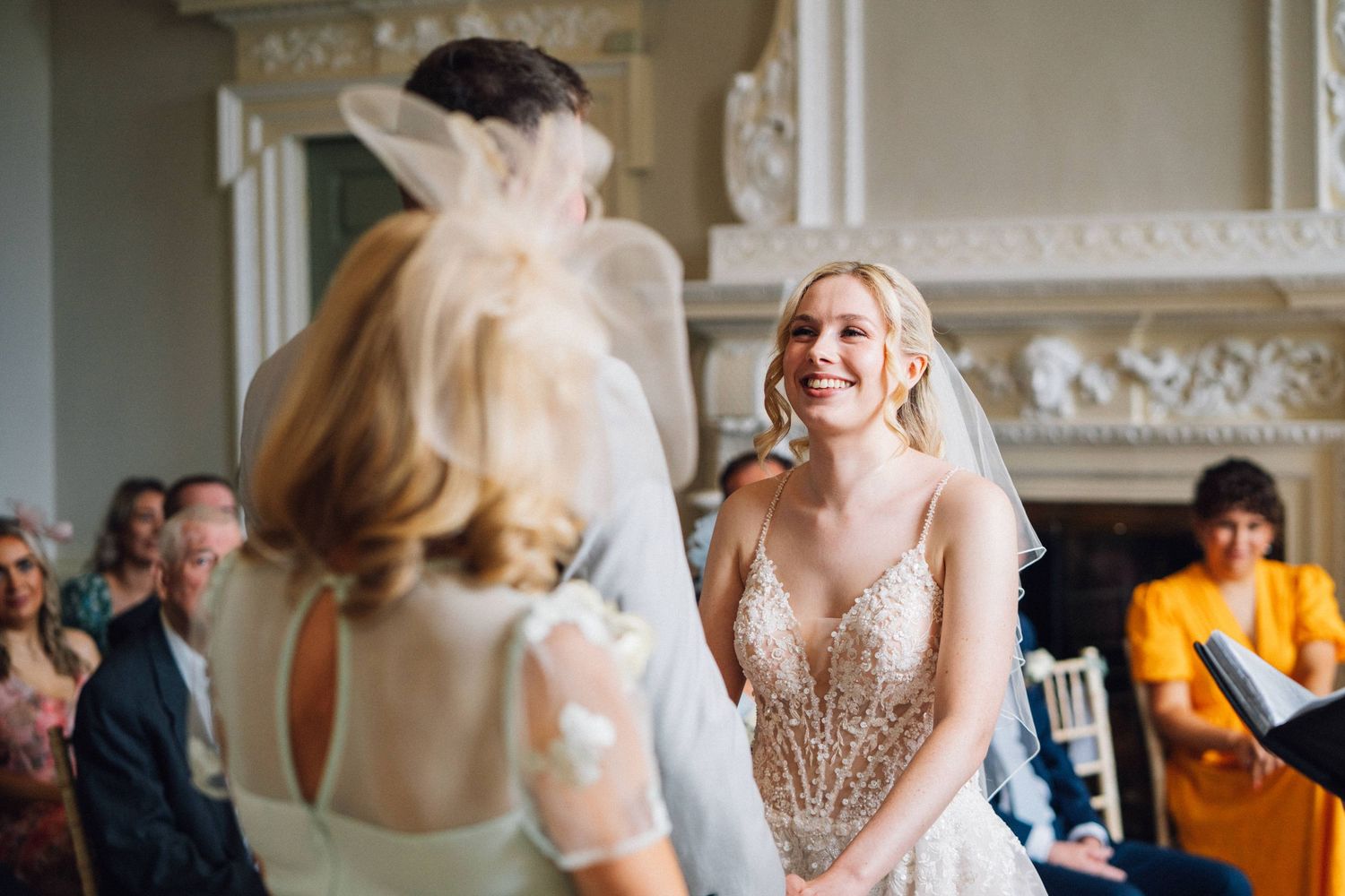 A bride and bridesmaids in blush pink dresses laugh together during a wedding celebration in an elegant room.