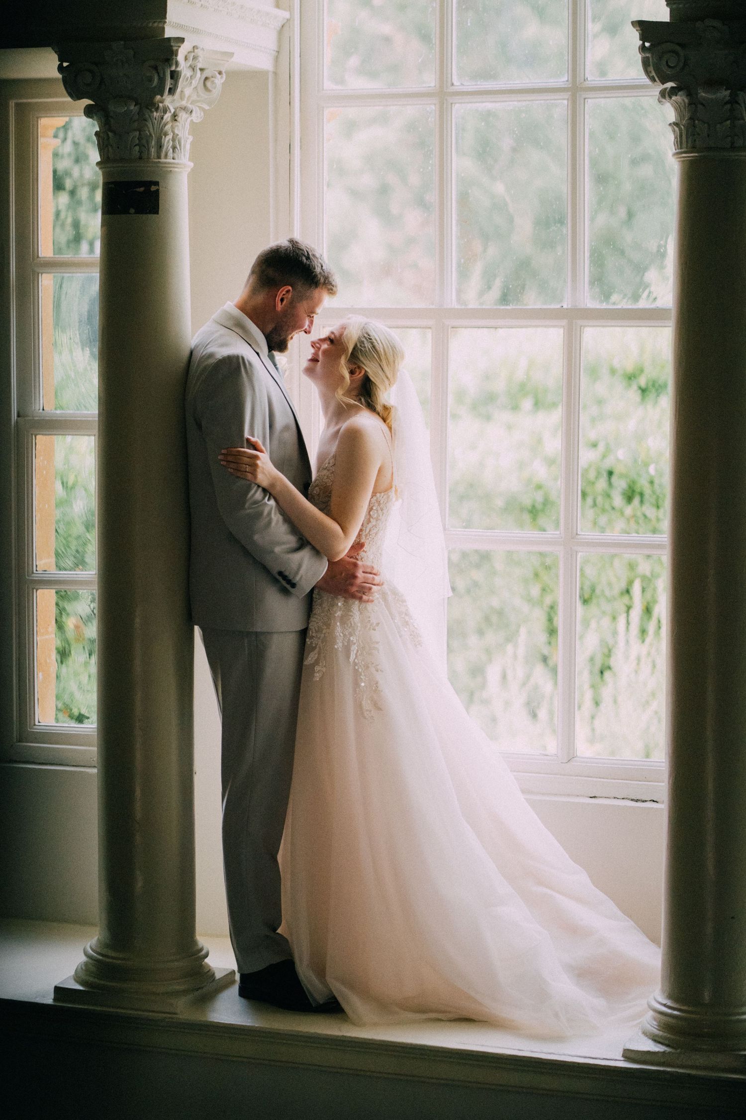 A romantic wedding portrait of a couple standing between elegant columns by a bright window.