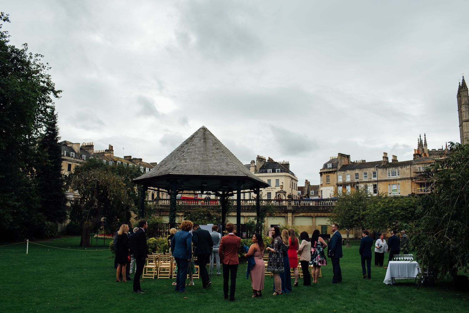 Wedding guests gather under a gazebo in an outdoor garden setting during an overcast day.