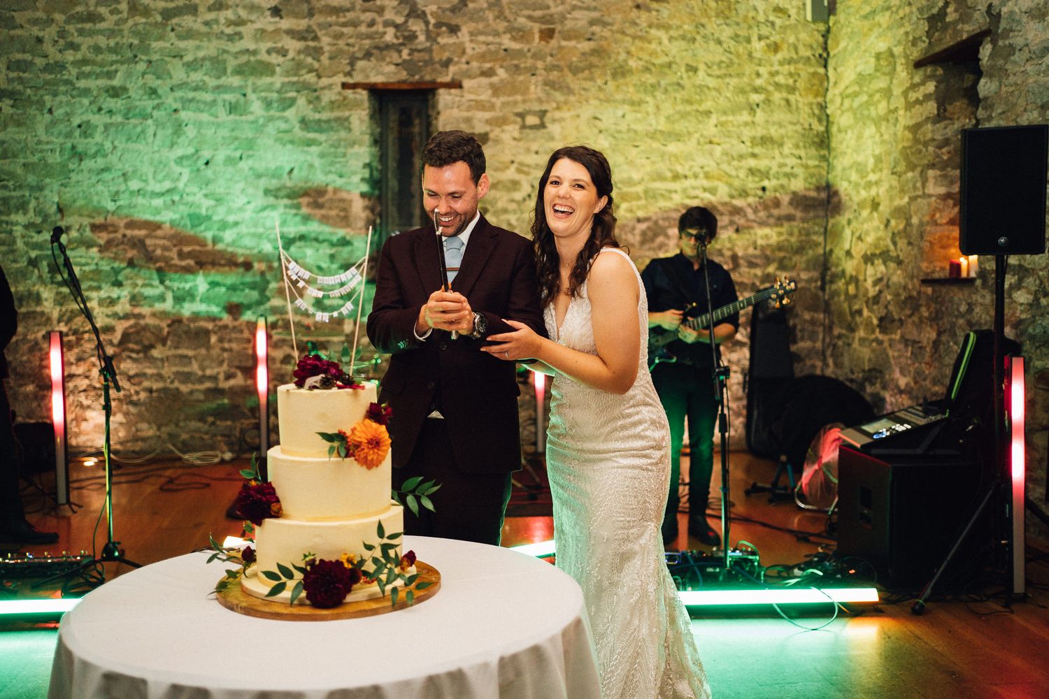 Wedding reception in a rustic brick venue with green uplighting and guests celebrating around a decorated cake table.