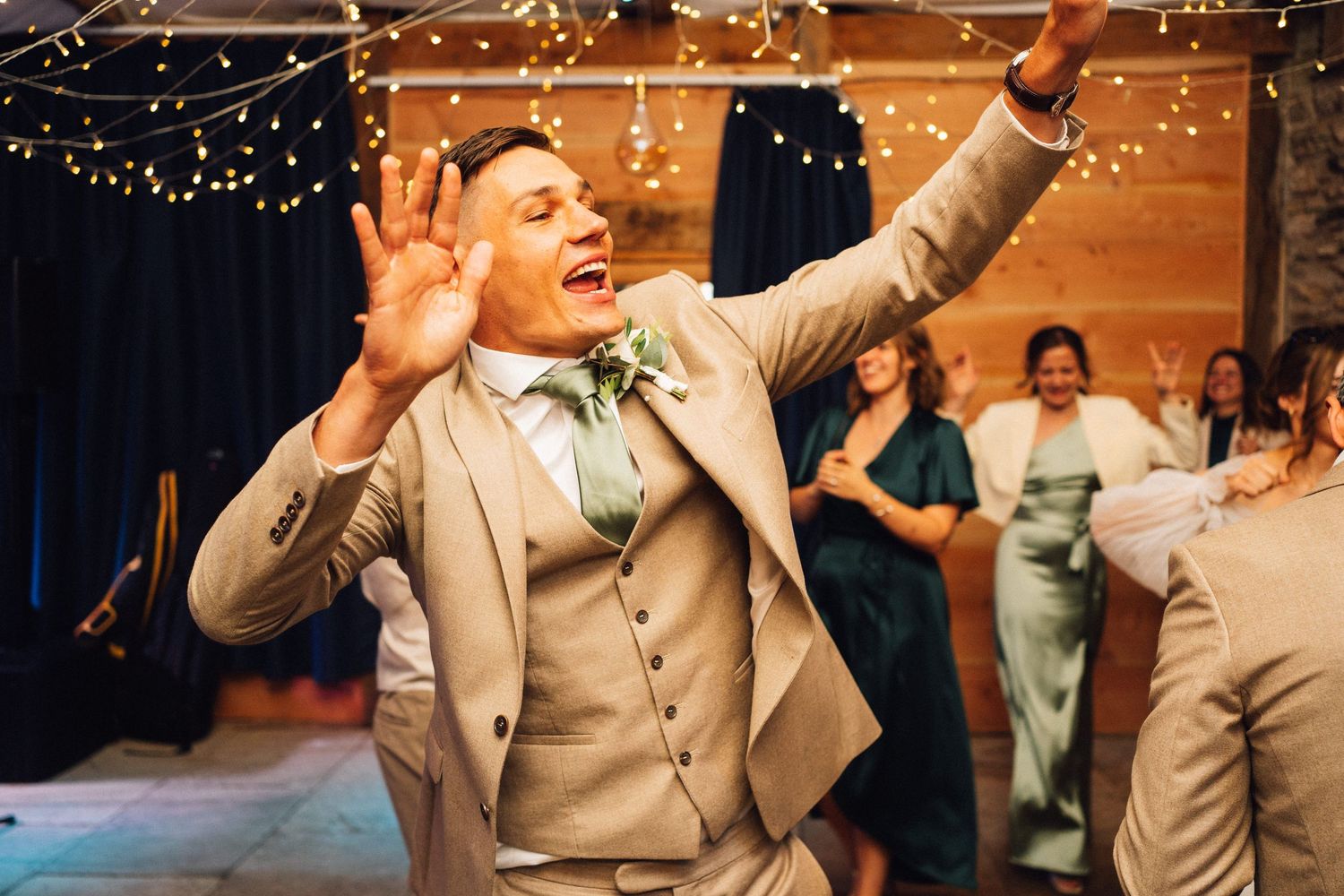 Wedding guests dancing and celebrating under warm string lights at an indoor reception venue.