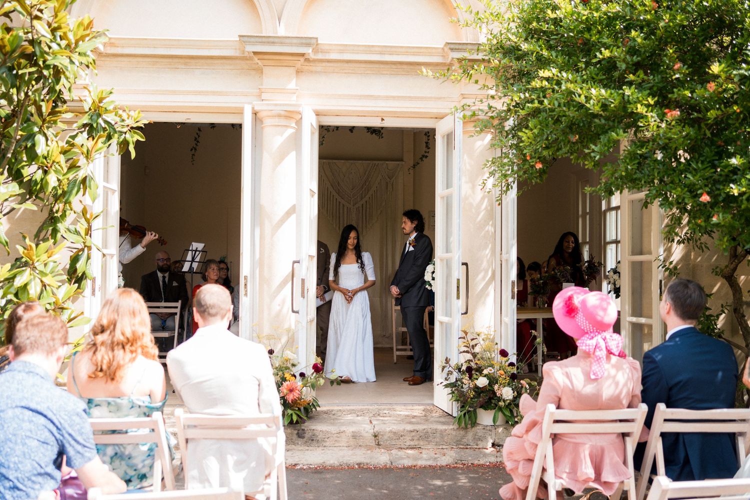 Outdoor wedding ceremony at elegant venue with bride and groom at entrance, guests seated in chairs amid floral arrangements.