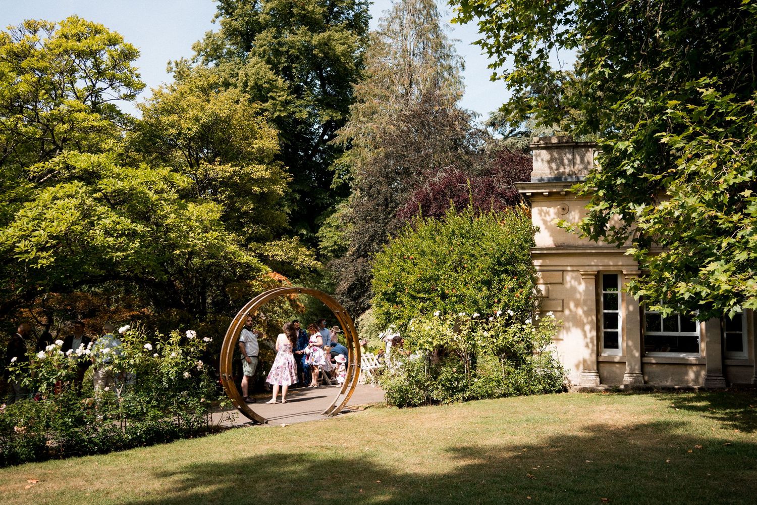 Wedding guests gather in a beautiful English garden with circular sculpture and stone manor house.