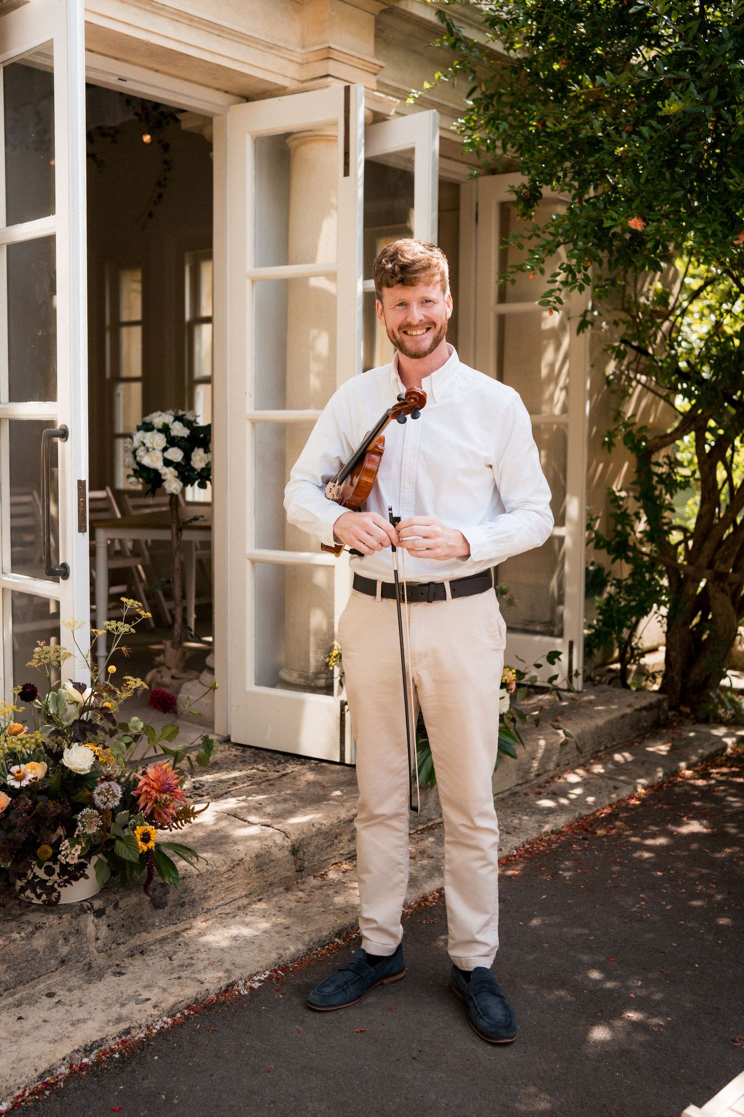 Smiling male violinist in white shirt and beige pants holding violin outdoors at elegant venue.