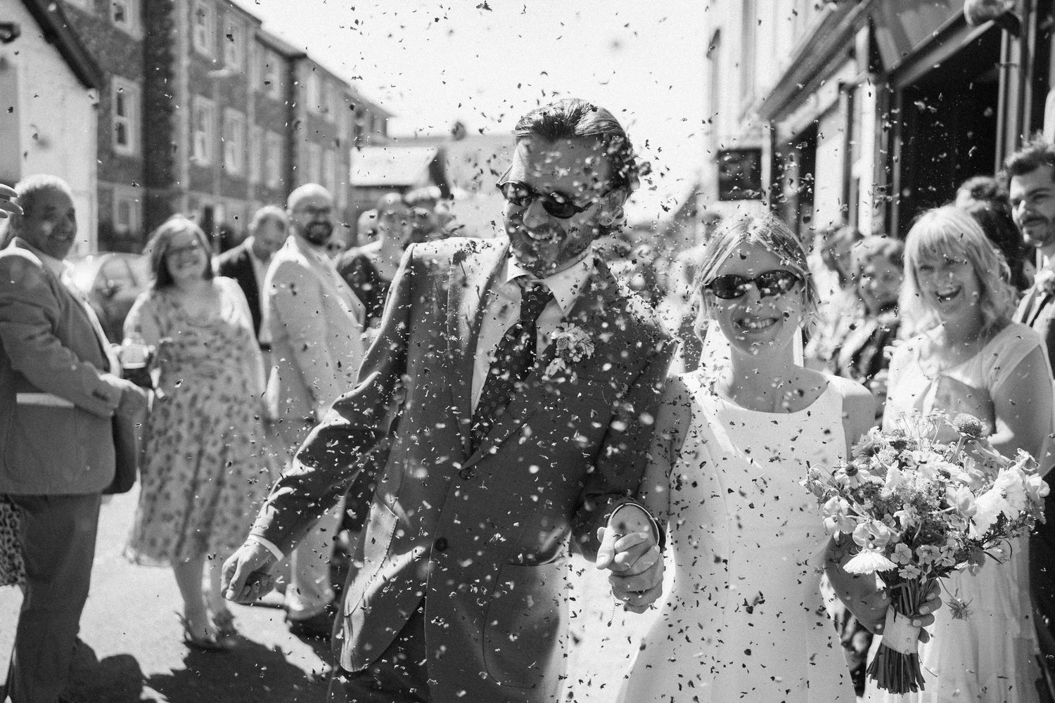 Bride and groom walk through confetti shower after wedding ceremony on sunny street.
