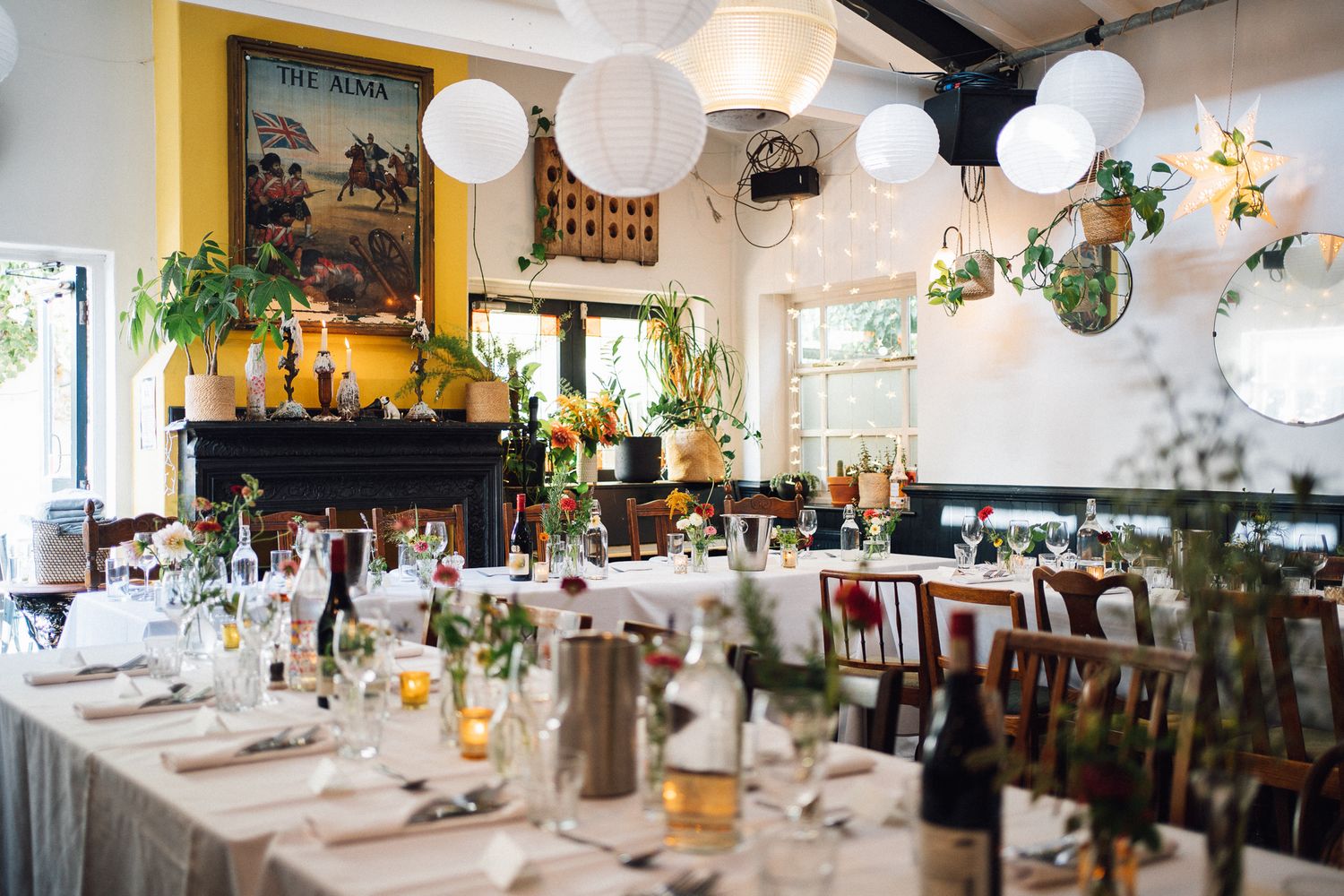 Elegant wedding reception venue with white paper lanterns, long dining tables, lush greenery, and a decorative fireplace.