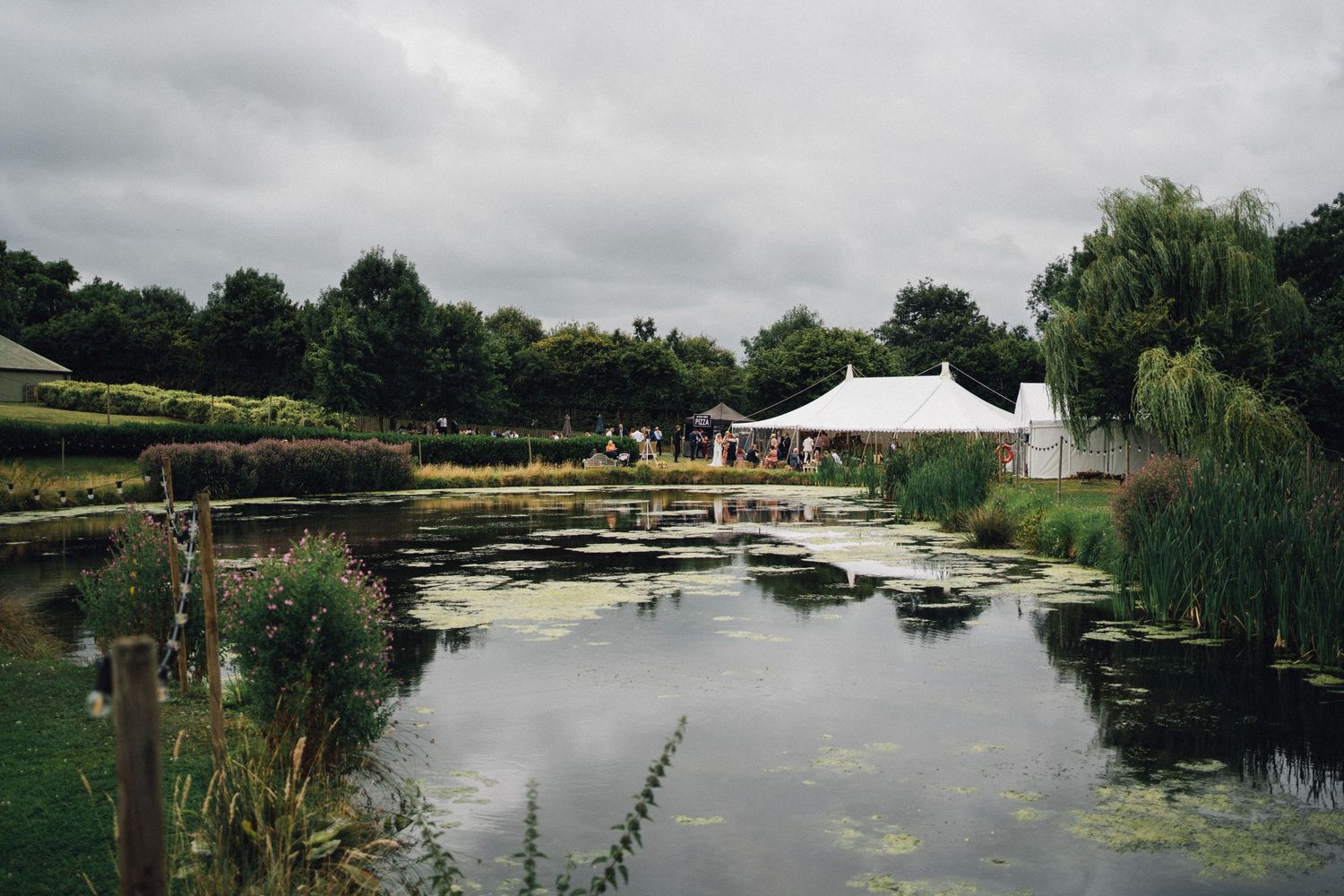 A serene garden pond with lily pads reflects a white marquee tent surrounded by lush trees under overcast skies.
