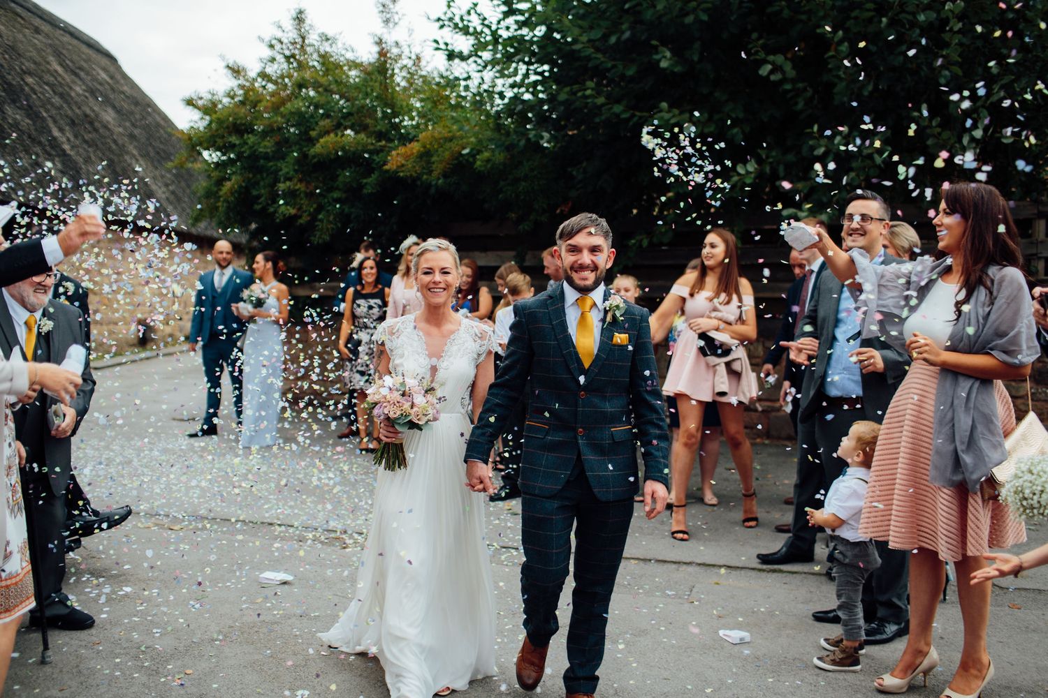 Panoramic view of wedding guests throwing confetti at a rustic outdoor venue.
