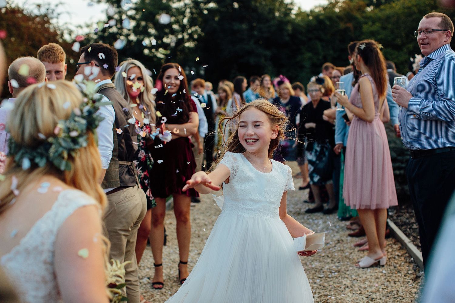 A young girl in a white dress twirls joyfully at an outdoor wedding celebration while guests watch and take photos.