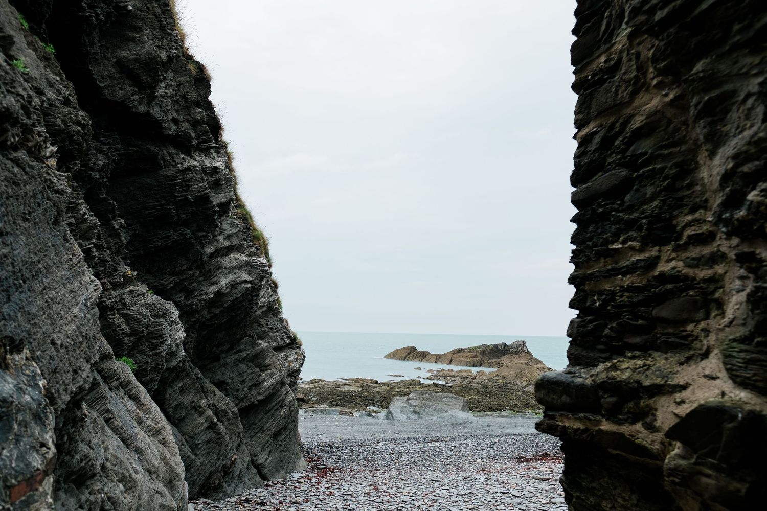 Dramatic rocky coastline with dark cliffs and pebble beach against cloudy sky.