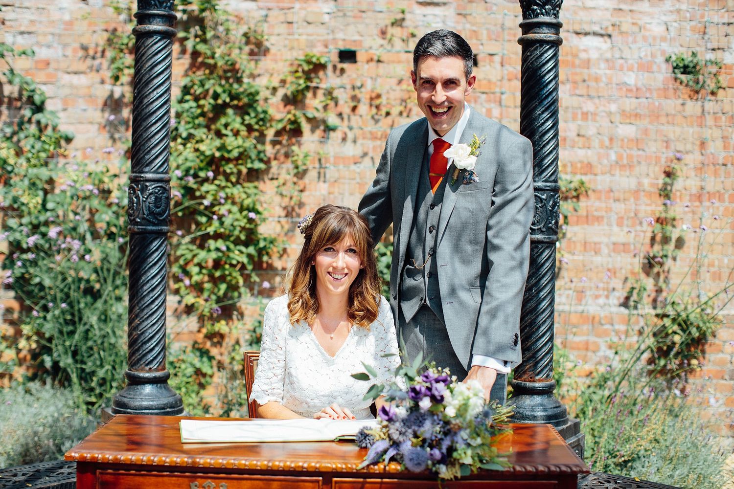 Couple participates in wedding ceremony under decorative columns with brick wall and greenery backdrop.