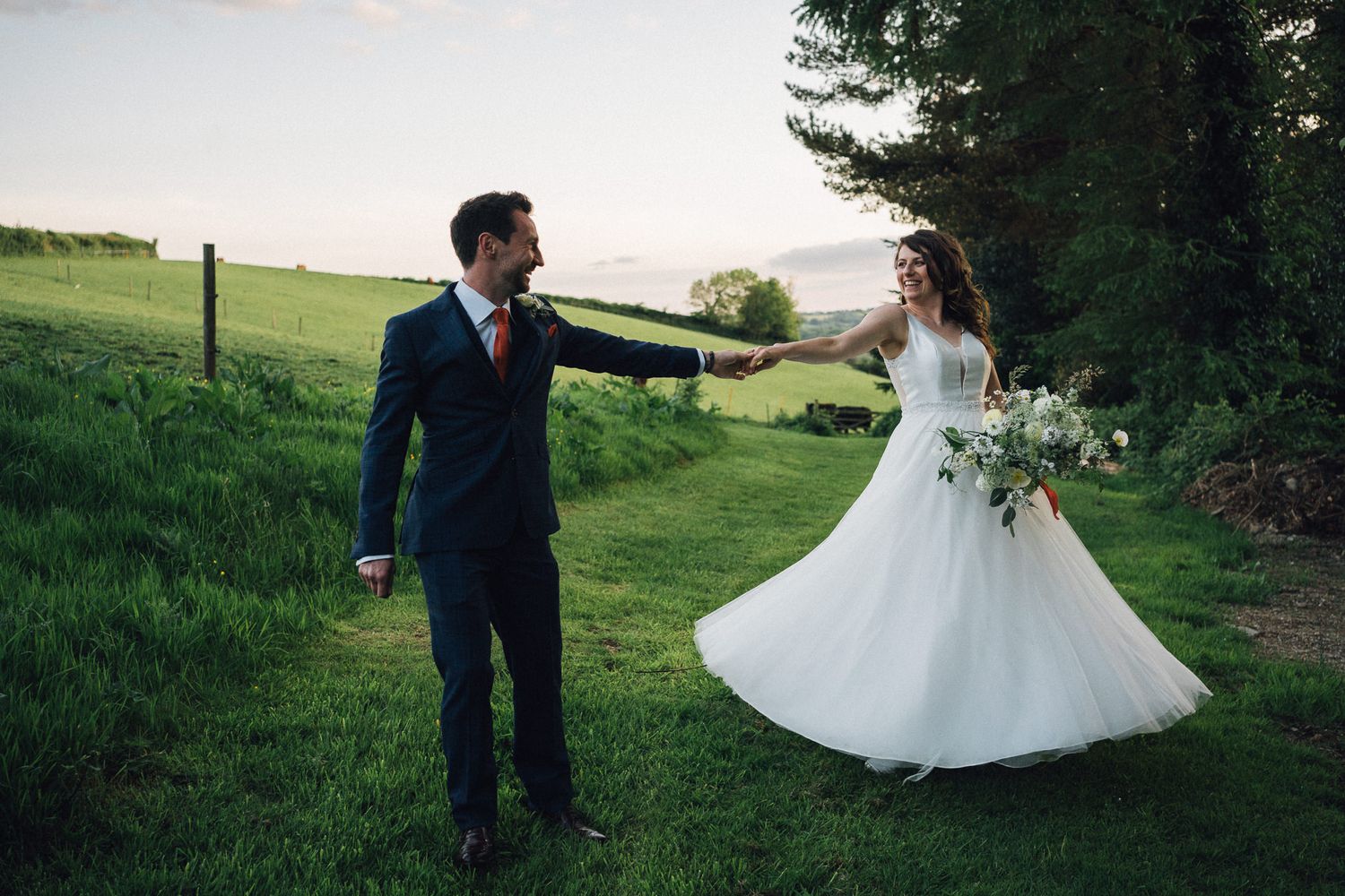 Bride's white dress flowing elegantly as couple walks through green meadow.