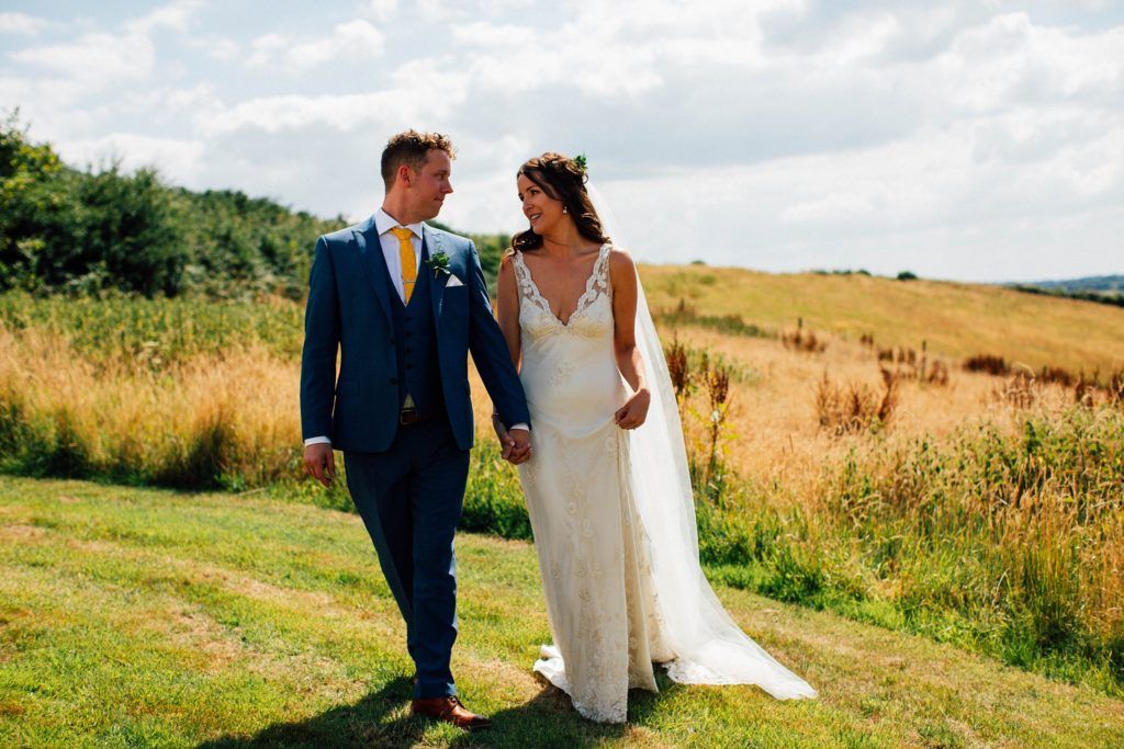 Newlyweds walk hand in hand through scenic countryside meadow.