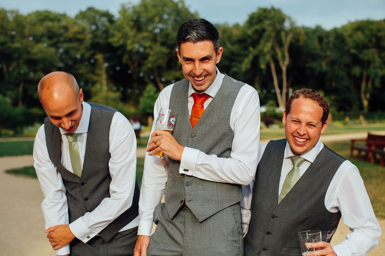 Three groomsmen in matching vests and ties share a candid laughing moment outdoors.