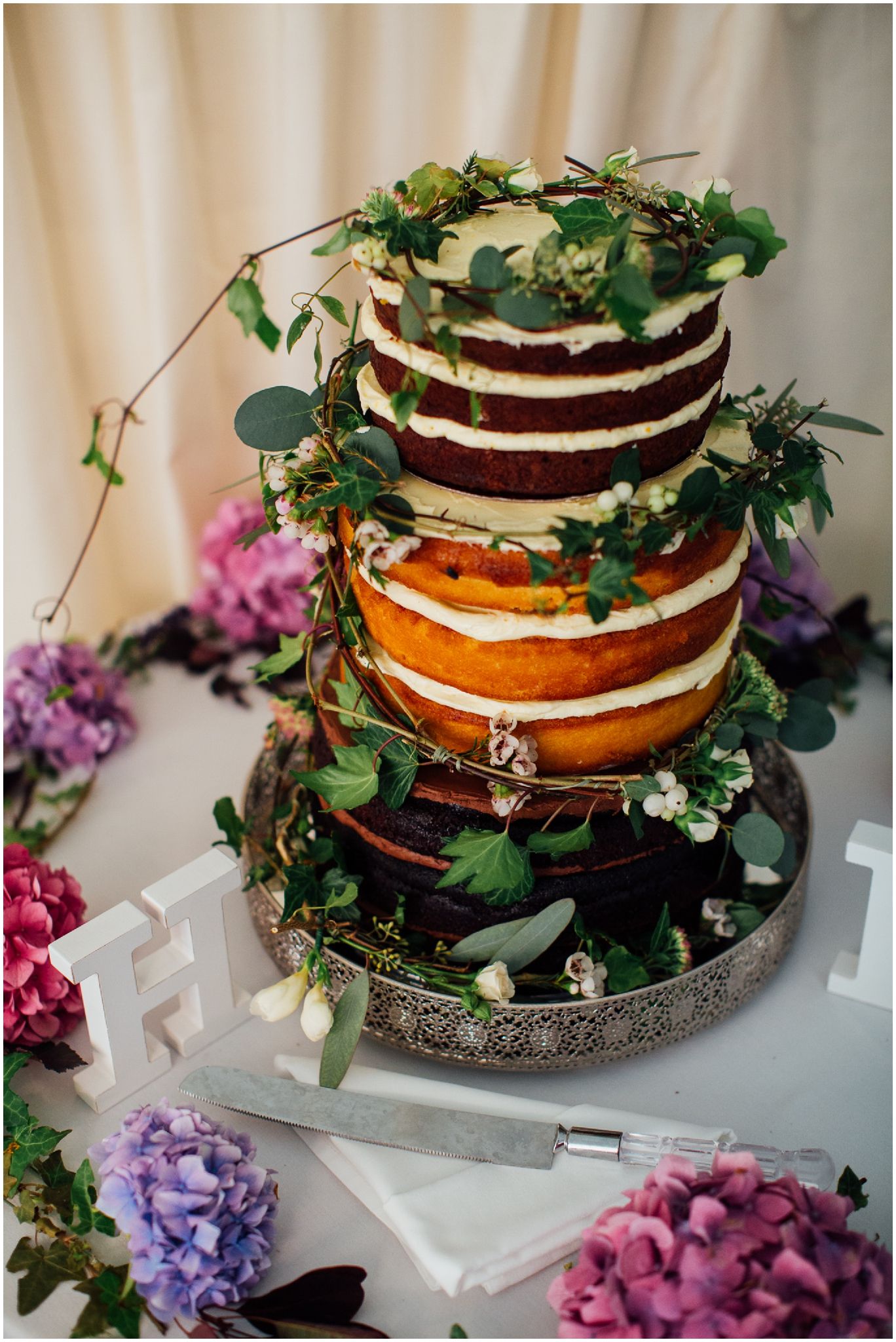 Rustic naked wedding cake decorated with greenery and purple flowers.