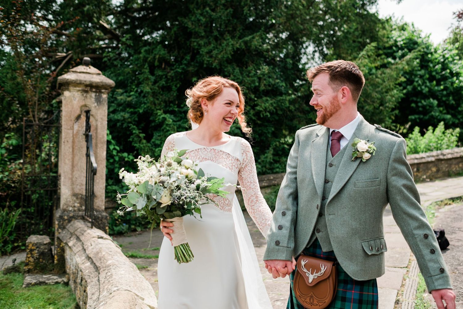 A joyful wedding couple in Scottish attire walking together on stone path surrounded by greenery.