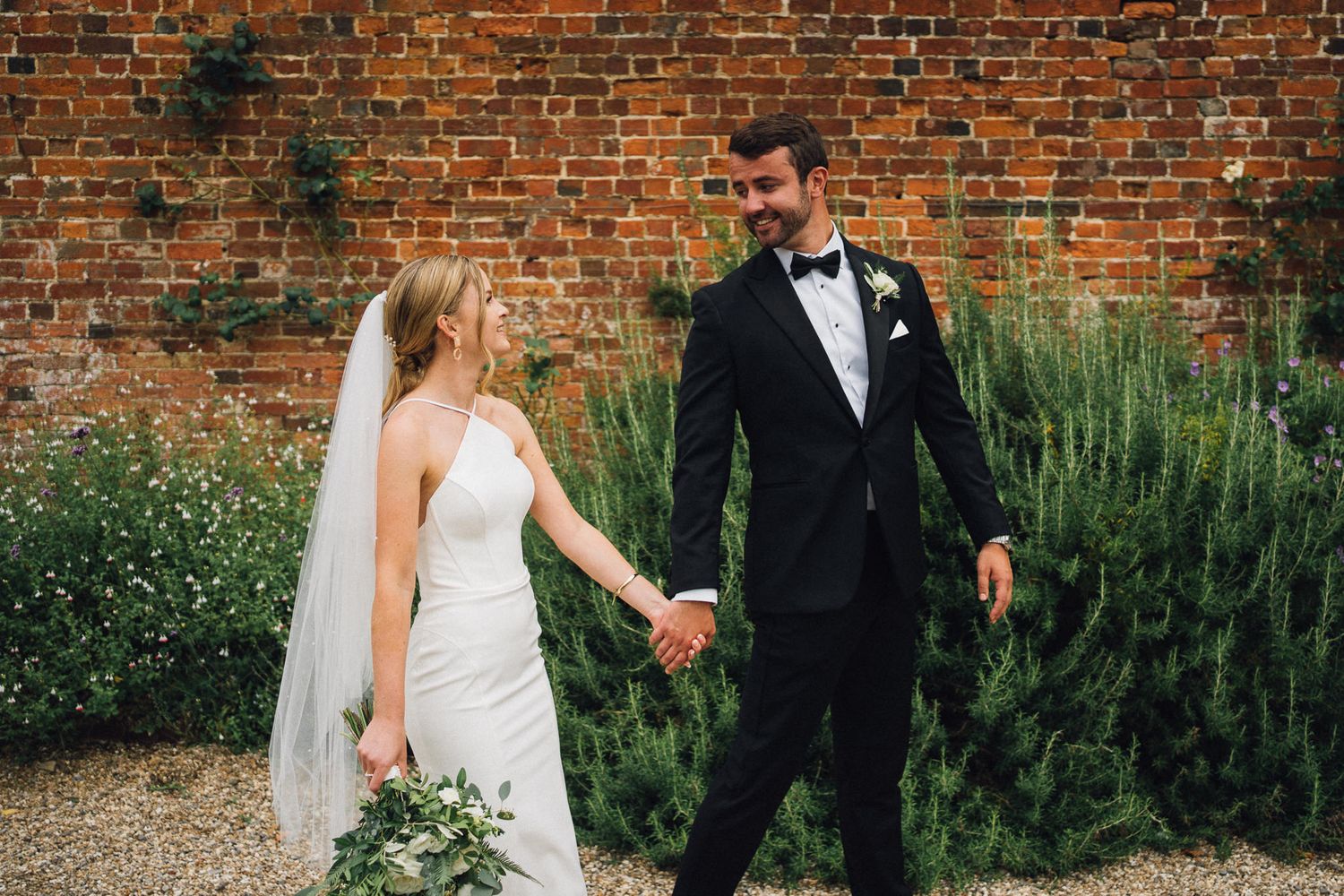A bride and groom walk together along a rustic brick wall with lush greenery in an outdoor garden setting.