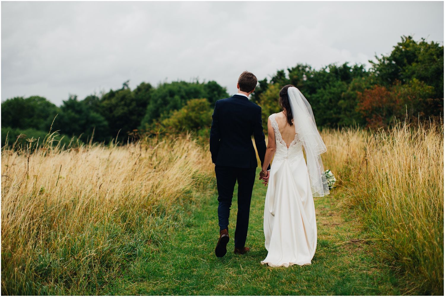 Wedding couple walks through tall grass field during outdoor photo session.
