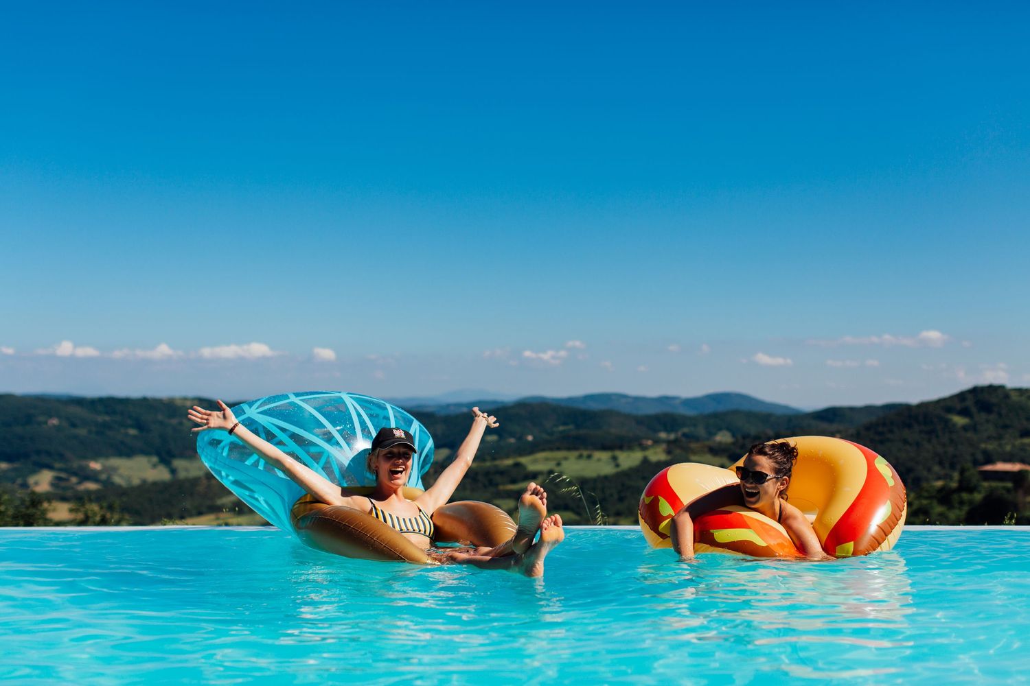 People floating on colorful pool floats in an infinity pool overlooking a scenic landscape.