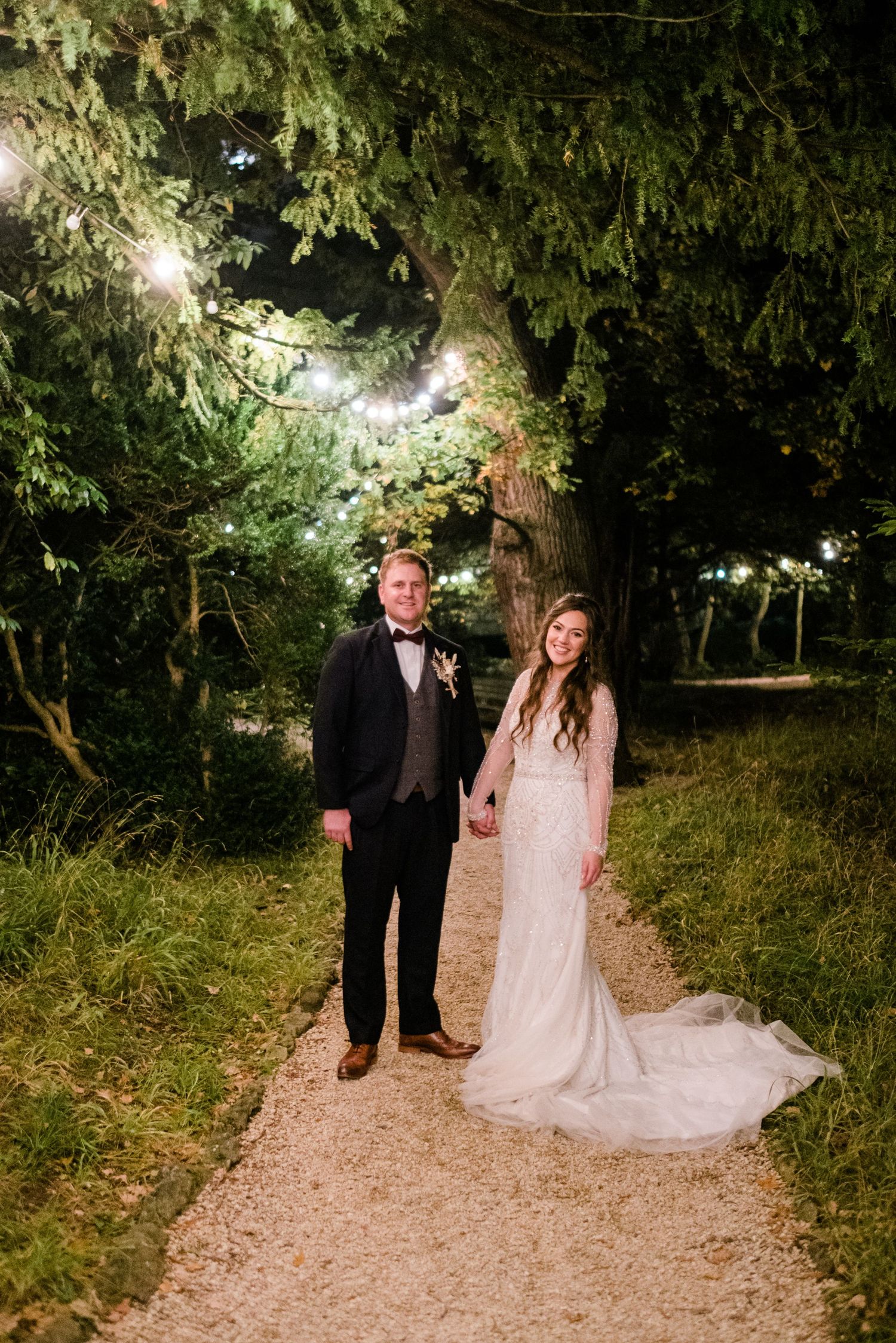 Romantic evening wedding photos on a tree-lined path with twinkling lights.