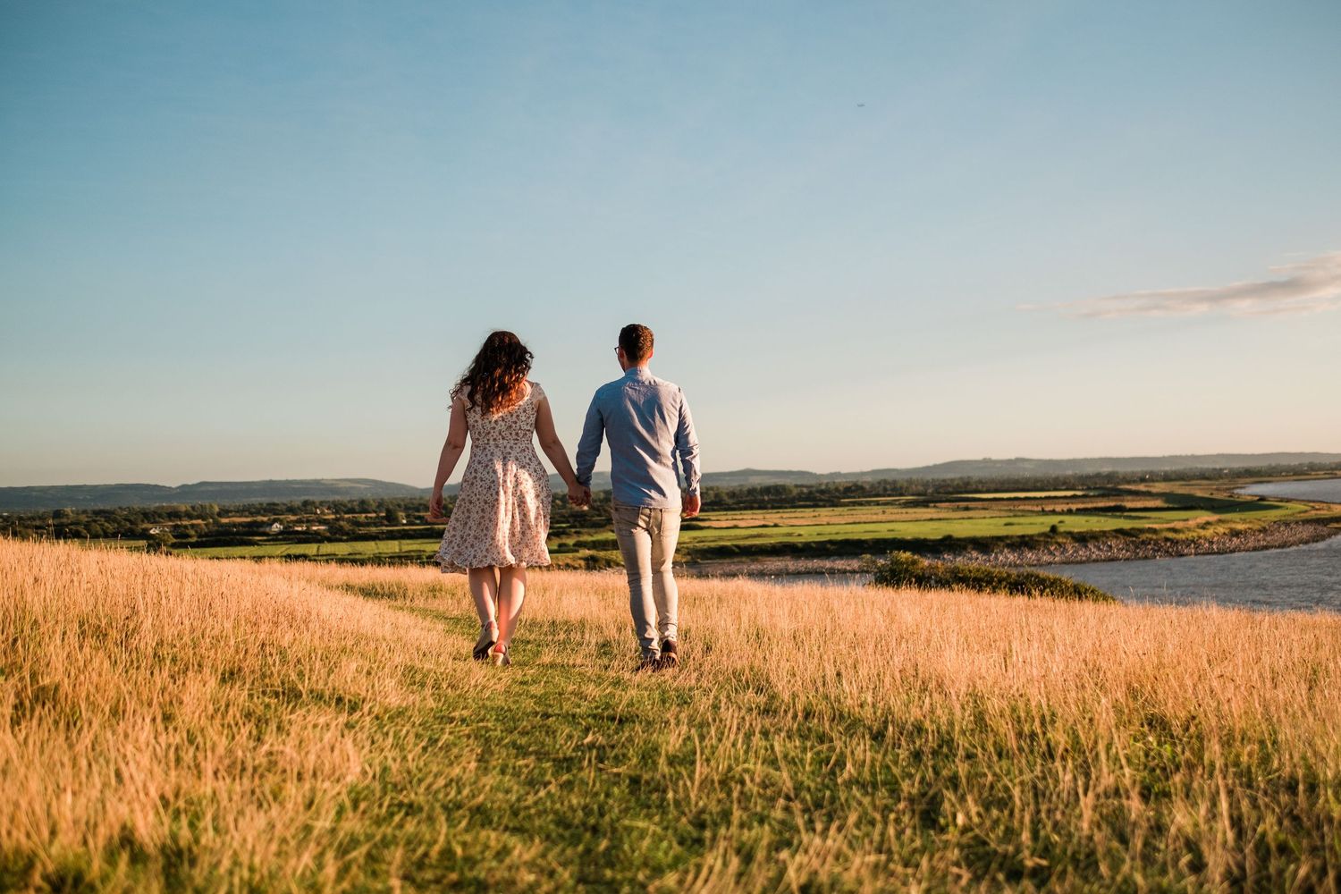 Couple walking hand in hand through golden wheat field overlooking coastal landscape at sunset.