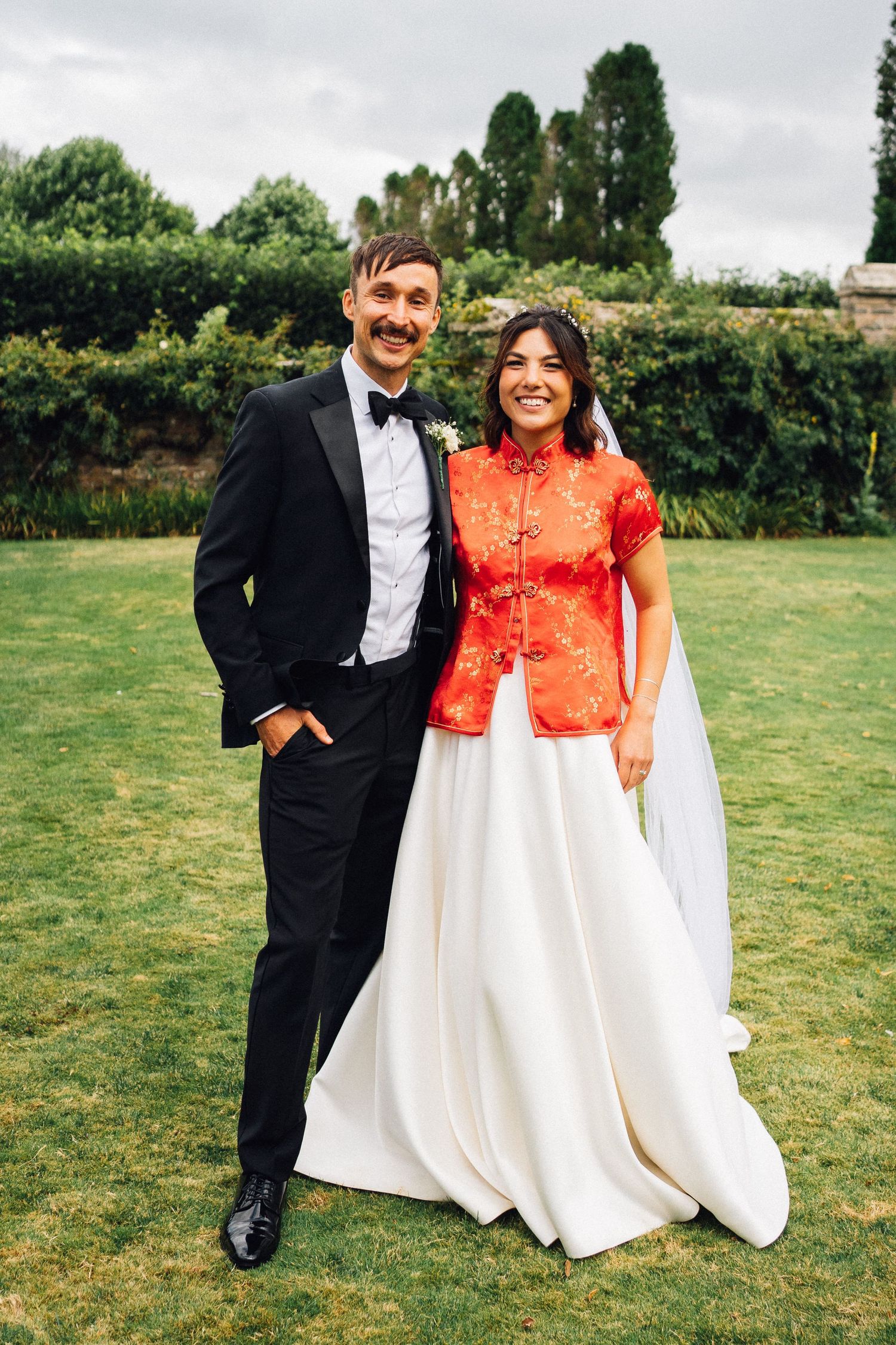 Wedding couple in formal attire with bride wearing white dress and red Chinese jacket outdoors on grass.