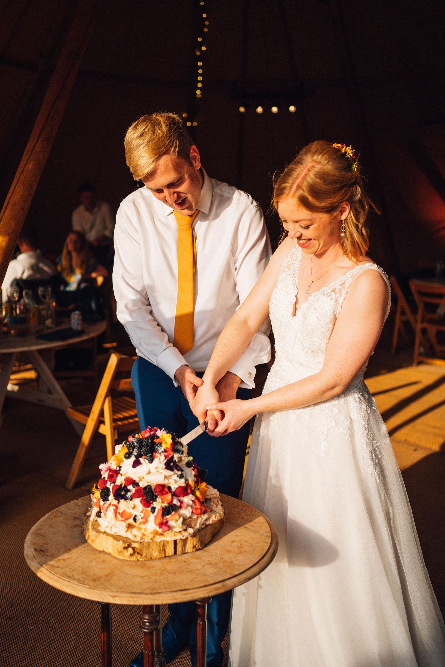 A couple cuts their rustic wedding cake adorned with fresh berries and flowers on a wooden serving board at their reception.
