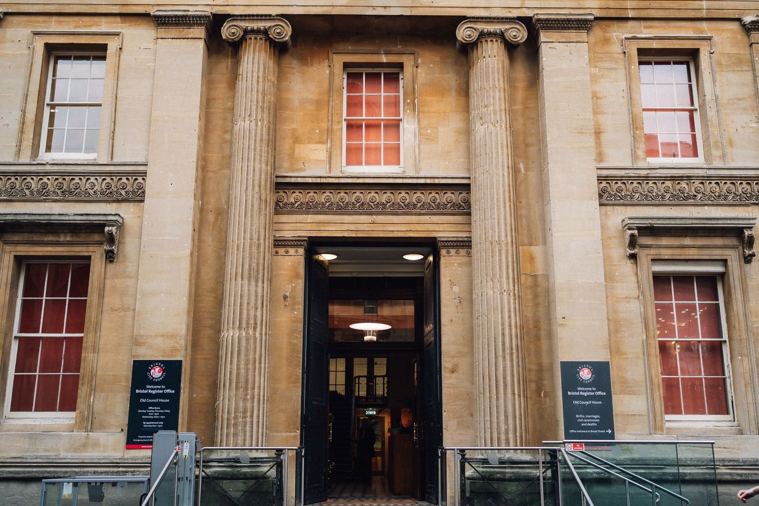Grand neoclassical building entrance with tall stone columns, red-framed windows, and open double doors.