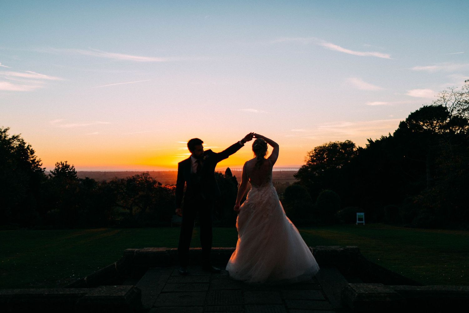 Silhouette of bride and groom dancing at sunset with vibrant orange sky and lush green trees in background.