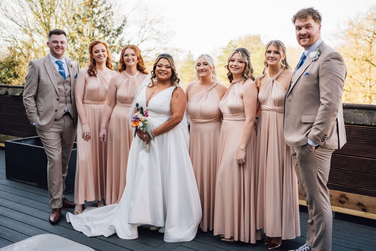 Wedding party in blush pink dresses and gray suits poses together outdoors during autumn celebration.
