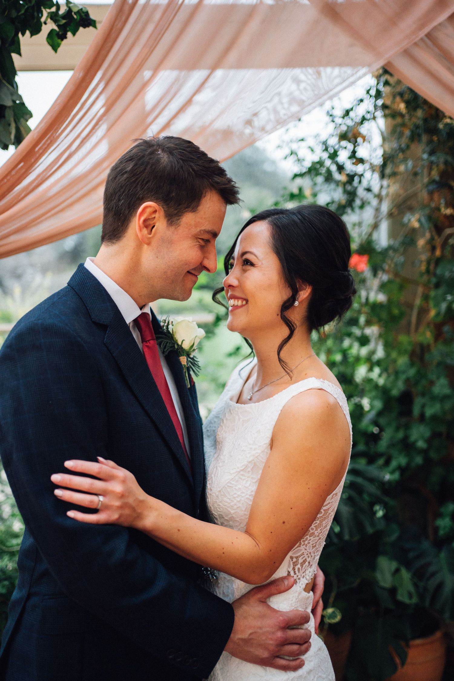 Newlywed couple sharing intimate moments under peach-colored drapes and greenery at their wedding.