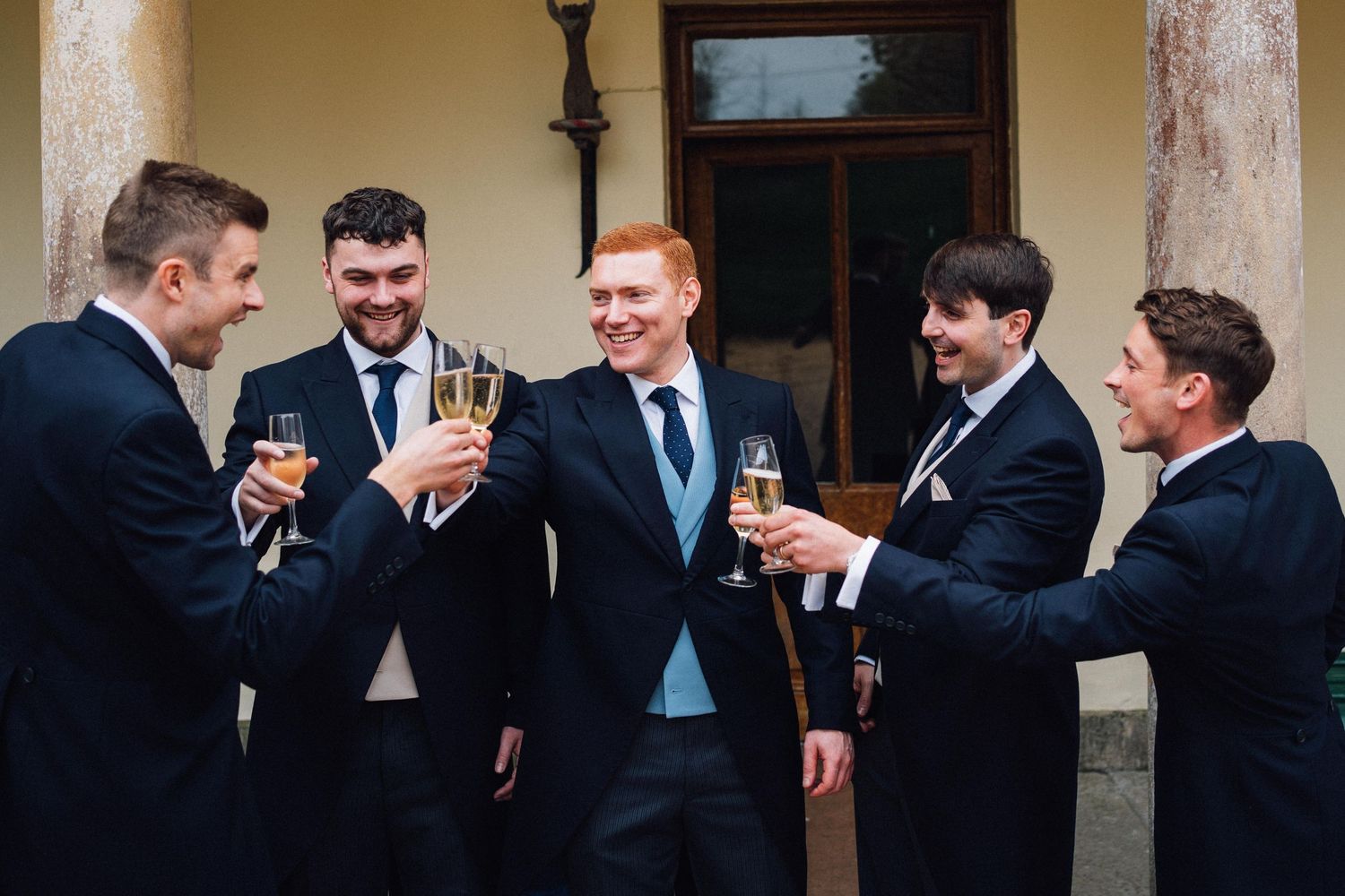 Groomsmen in dark suits sharing drinks and laughs during wedding celebrations.