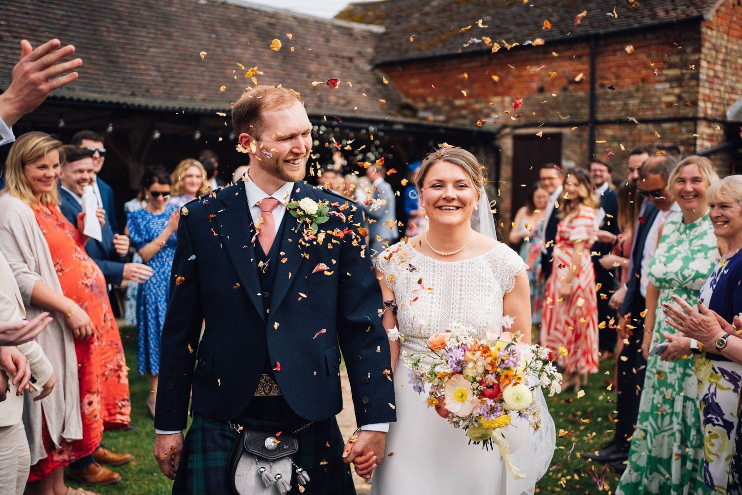 Wedding party and guests celebrating with confetti outside a rustic venue.