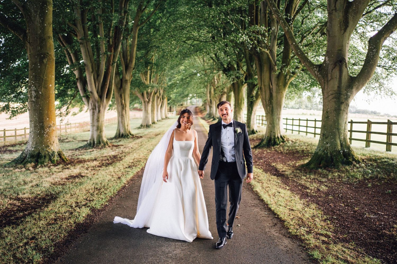 Romantic walk through a lush green tree-lined avenue during a wedding photoshoot.