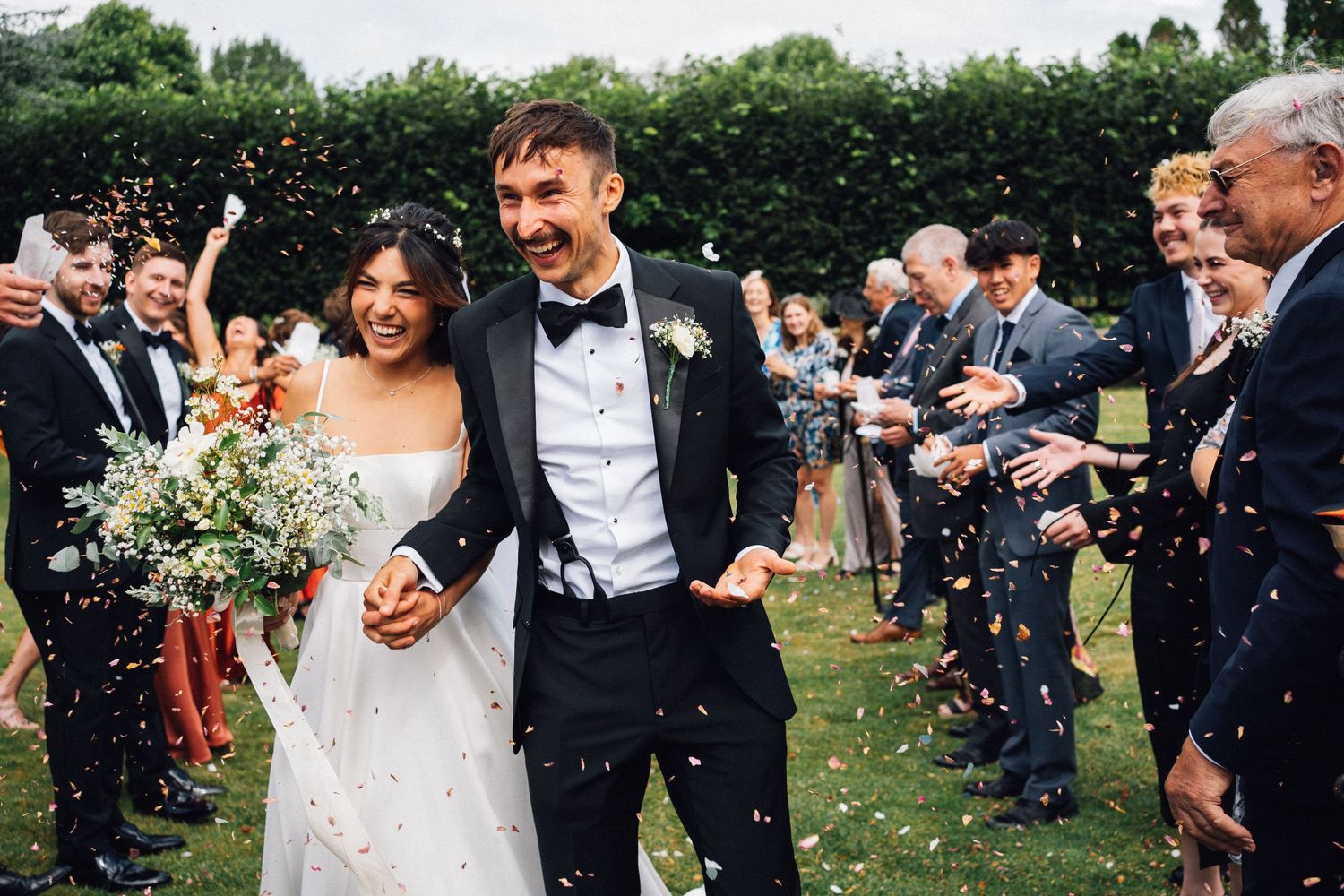 Wedding guests throw confetti as newlyweds celebrate during an outdoor garden ceremony.