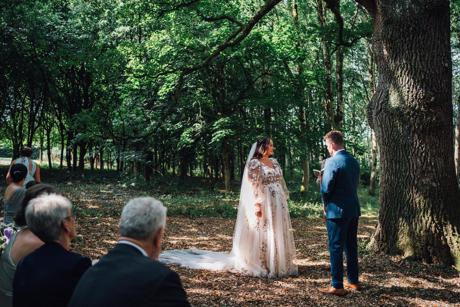 A wedding ceremony takes place in a serene forest setting with natural light streaming through trees.