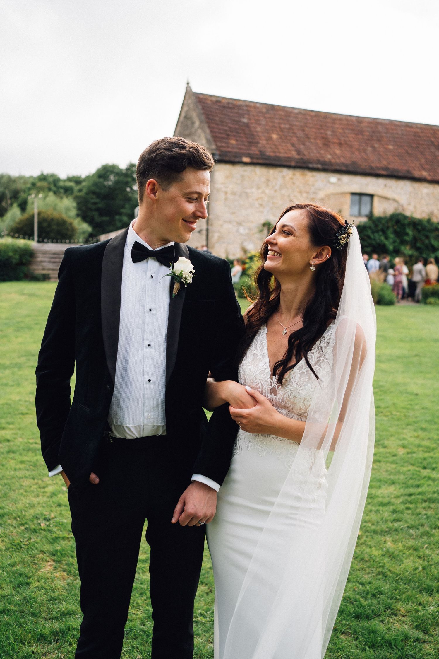 A joyful wedding couple shares candid laughter on a grassy lawn with stone buildings in the background.