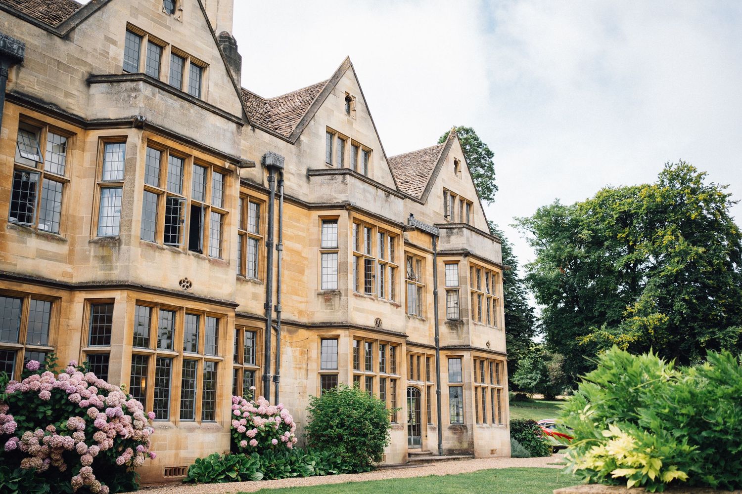 Historic English manor house with honey-colored stone walls, bay windows, and blooming pink hydrangeas in the garden.